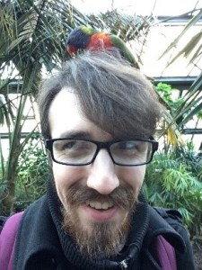 Picture of a man with brown hair, beard and glasses with a parrot on his head