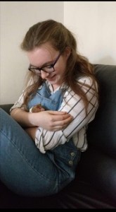 Young woman with long light brown hair wearing denim dungarees holding a hamster and looking at it