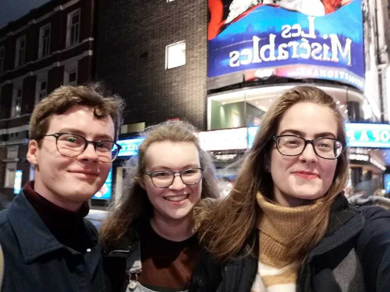 Two young brown haired women and a brown haired boy in glasses outside a theatre with the sign "Les Miserables"