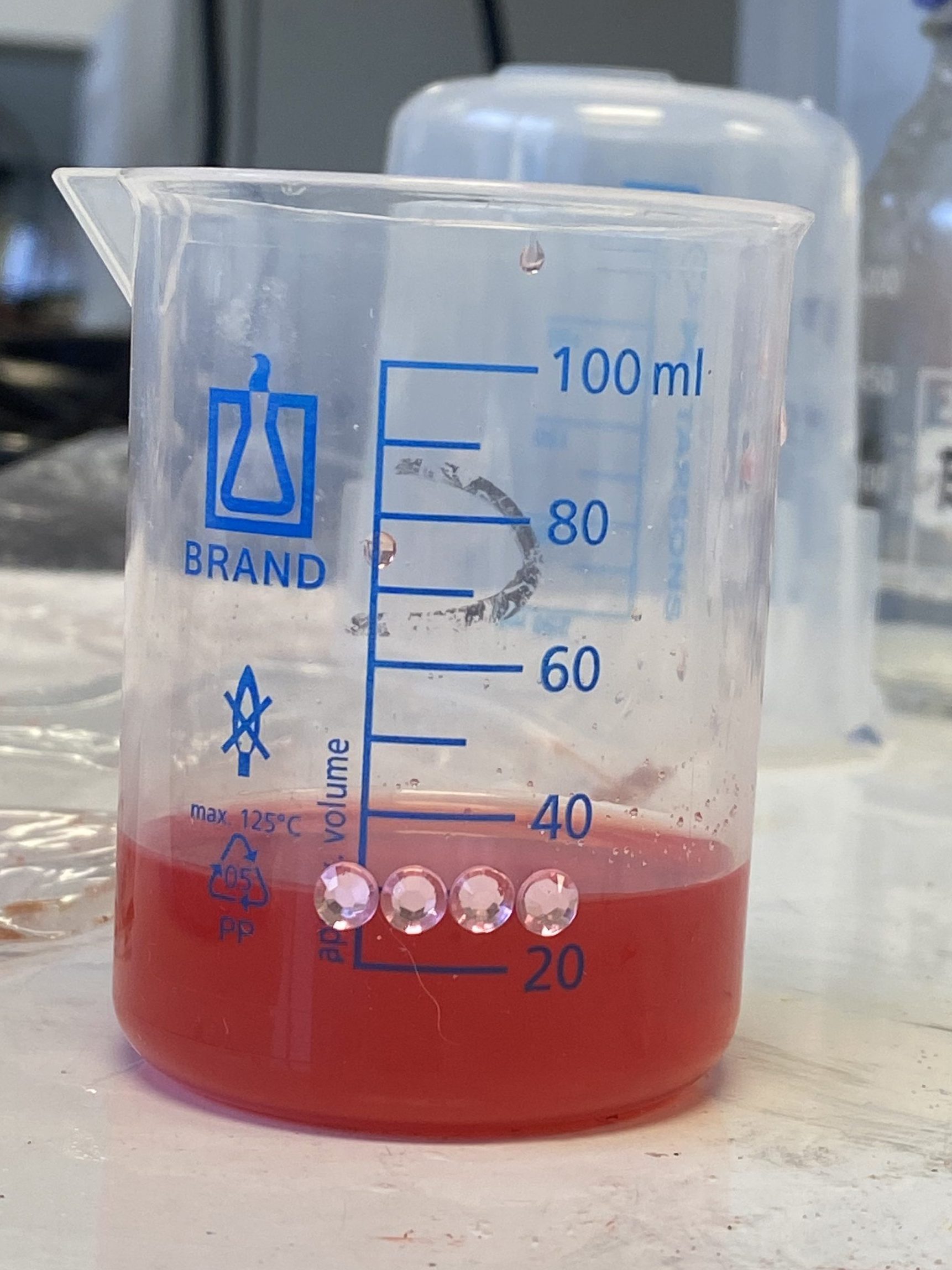 The image shows a 100mL plastic beaker, filled to 25mL with the dyed salt water solution. There are gems stuck onto the beaker at the 25mL level.